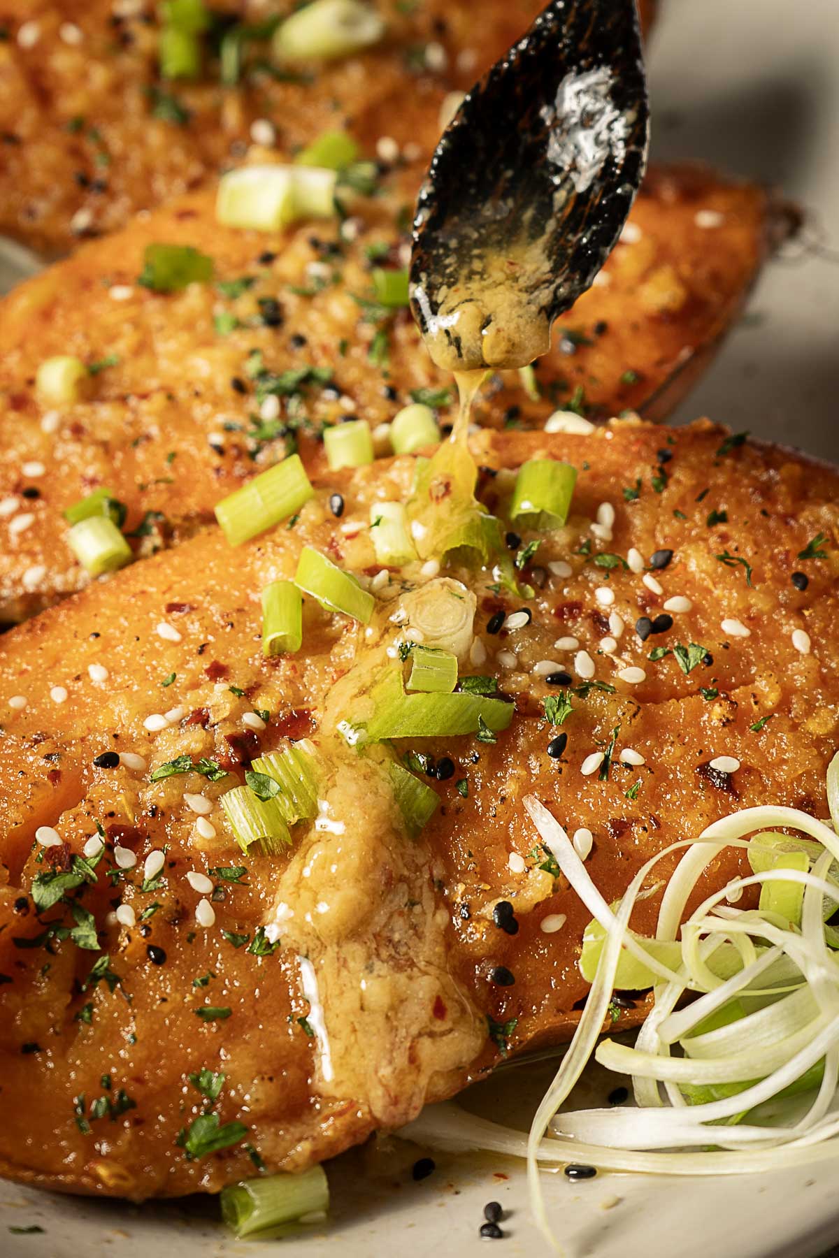 Close-up of three pieces of seasoned baked salmon topped with chopped green onions, sesame seeds, and spices, with a spoon drizzling sauce over one piece.