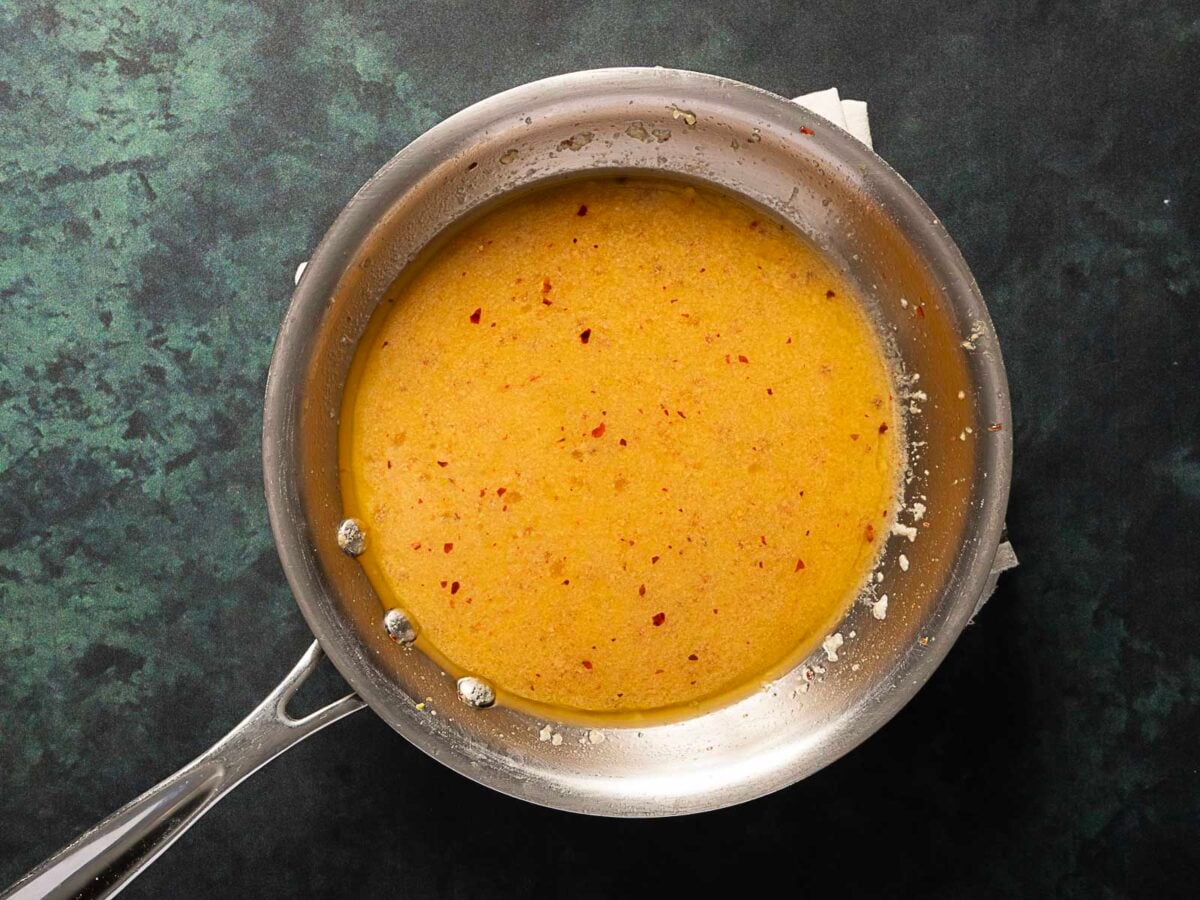A stainless steel pan containing a yellow-orange sauce with red pepper flakes sits on a dark green countertop.