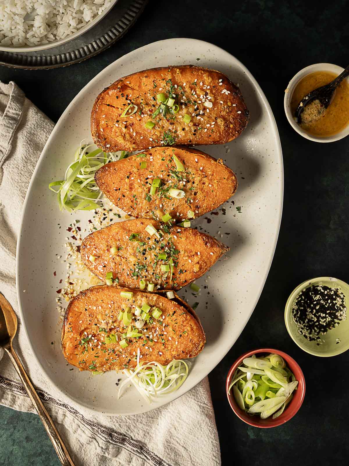 Four halves of roasted sweet potatoes garnished with sesame seeds and chopped green onions on an oval plate, surrounded by small bowls of sauces and toppings.