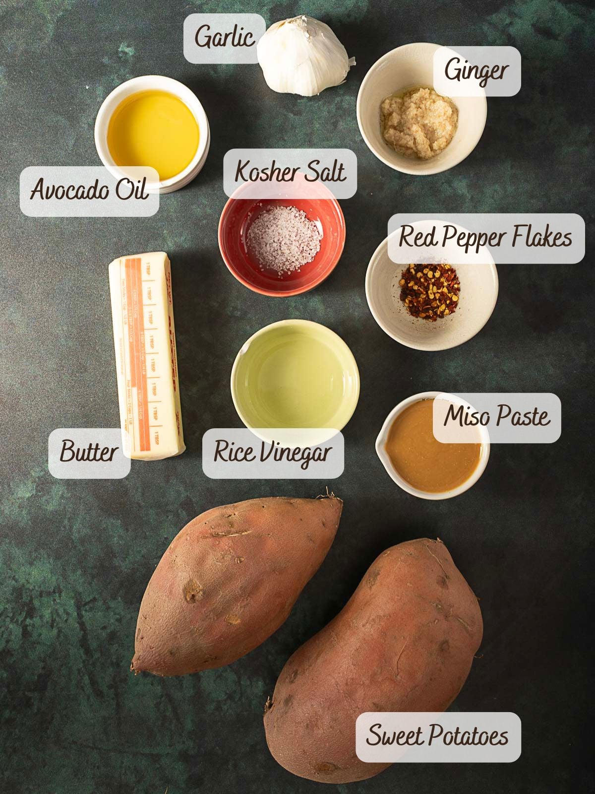Ingredients including garlic, ginger, avocado oil, kosher salt, red pepper flakes, butter, rice vinegar, miso paste, and two sweet potatoes arranged on a dark surface—perfect for preparing a flavorful miso sweet potato recipe.