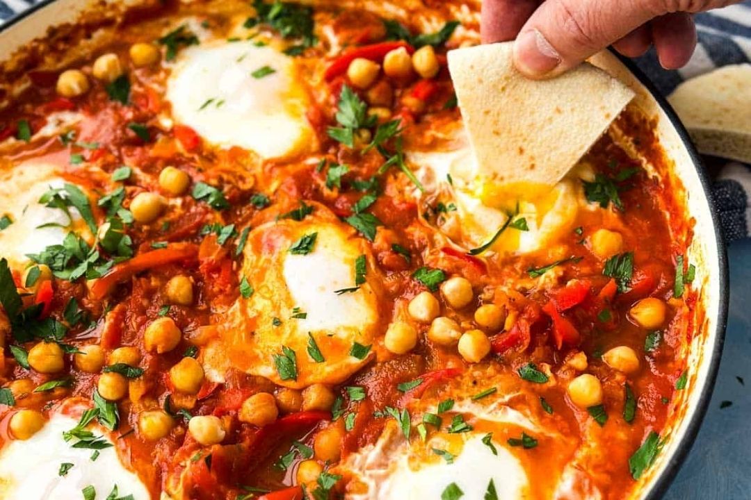 A hand dips pita bread into a skillet of shakshuka with poached eggs, chickpeas, tomatoes, peppers, and herbs—a vibrant dish perfect for those who love savory breakfasts and are seeking new breakfast recipes to try.