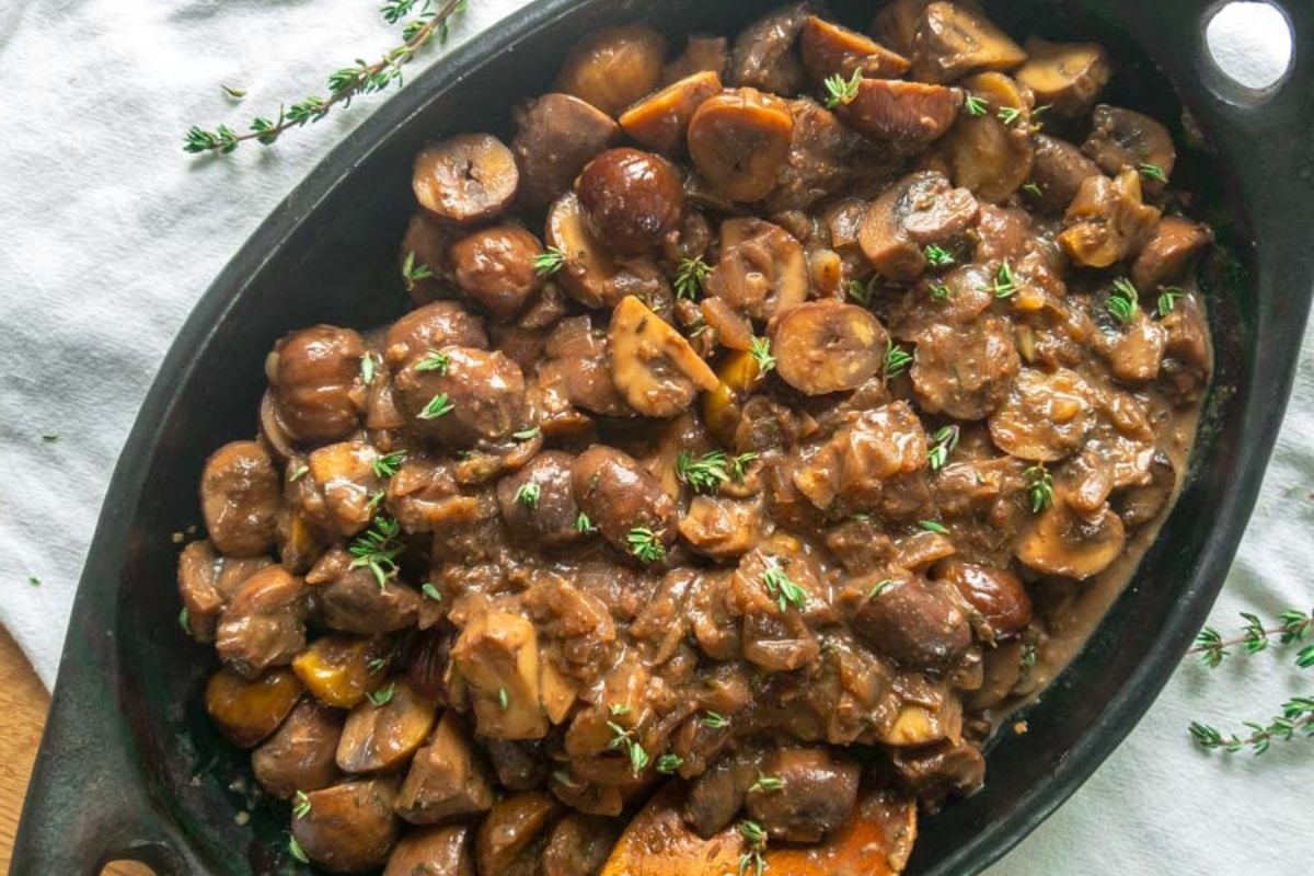 A black dish filled with cooked mushrooms in a flavor-packed brown sauce, garnished with fresh thyme sprigs, sits on a white cloth—making it an irresistible addition to your collection of side dishes.