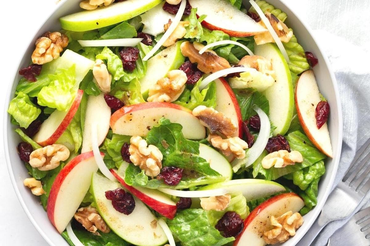 Bowl of salad with sliced apples, walnuts, dried cranberries, romaine lettuce, and onion strips, seen from above—perfect for fall salads and celebrating salad season.