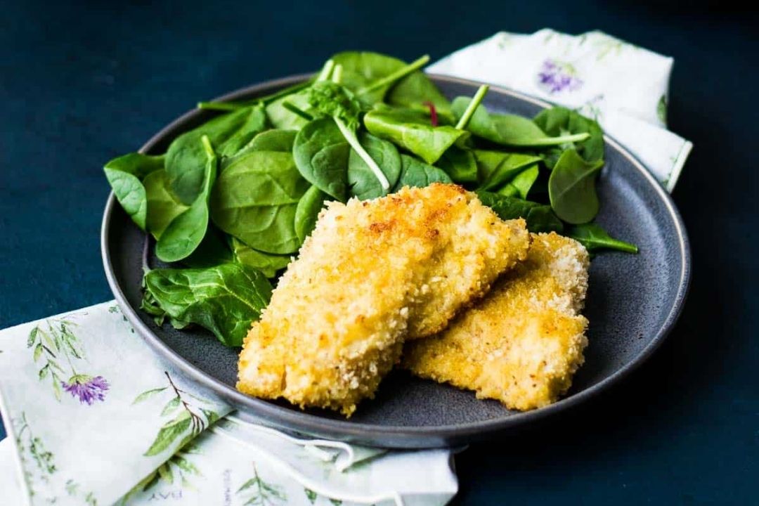 A gray plate with two breaded, baked fish fillets and a serving of fresh spinach leaves, set on a floral napkin—perfect inspiration for those seeking easy fish recipes or searching for the best fish dishes to enjoy at home.