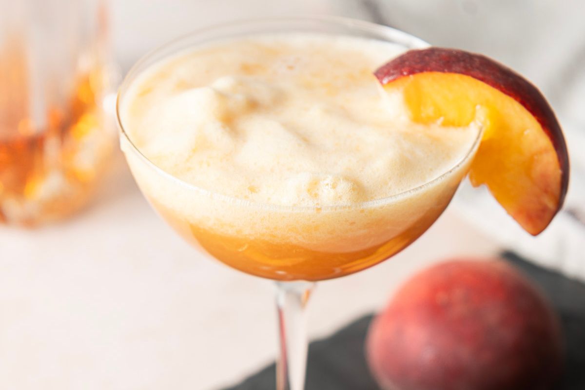 A cocktail glass filled with a frothy orange drink, garnished with a peach slice on the rim; a whole peach is nearby—perfect inspiration for your next round of brunch cocktails.