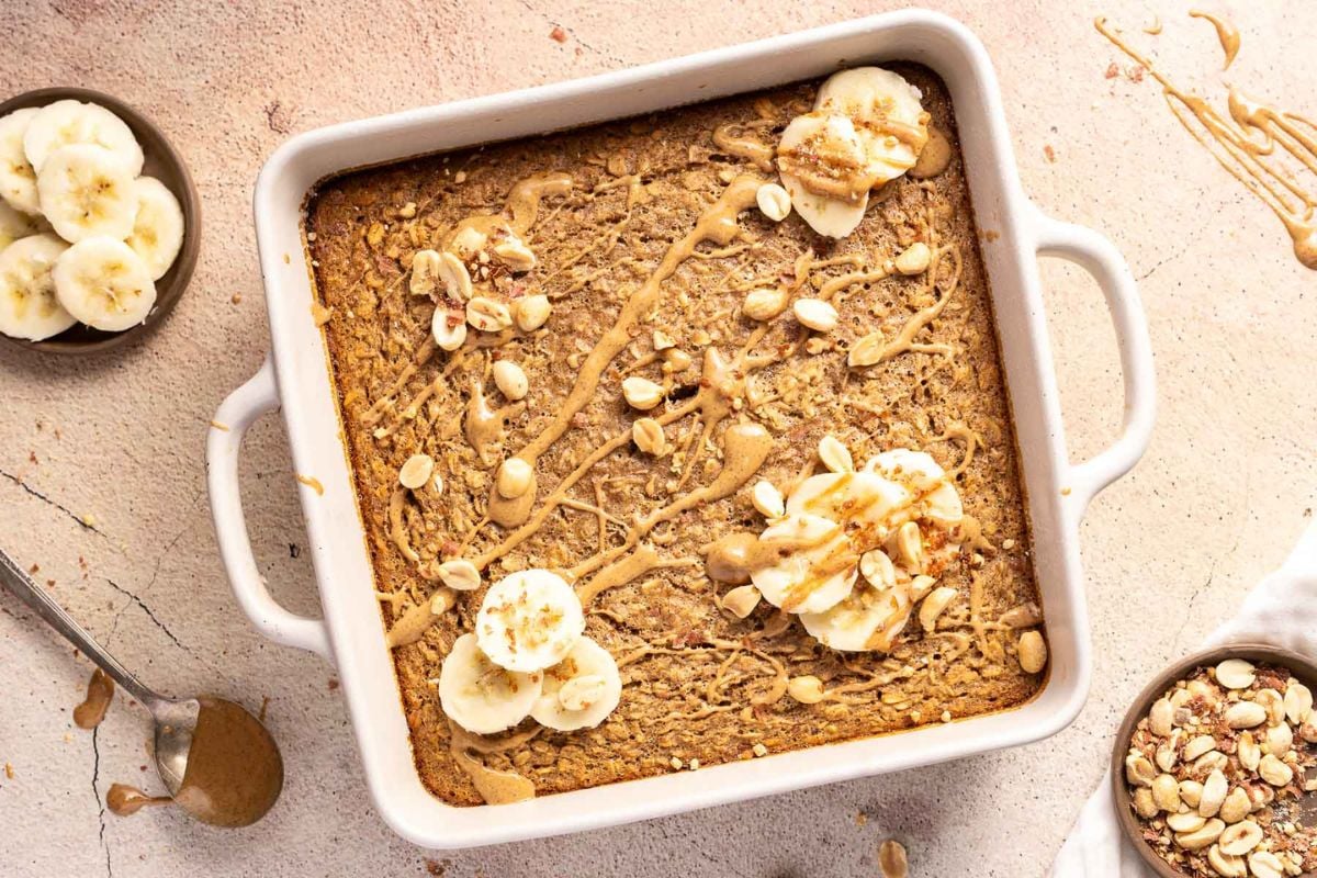 A baked oatmeal dish topped with sliced bananas, chopped nuts, and a drizzle of nut butter, beside a small bowl of banana slices and a spoon with nut butter—this wholesome option is perfect for healthy eating and makes a delicious addition to your family meals.