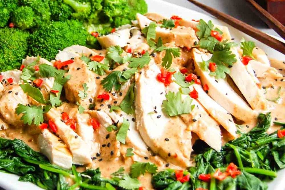 Sliced rotisserie chicken breast with sauce, garnished with cilantro, sesame seeds, and red chili, served on spinach with broccoli—a perfect choice for easy meals.