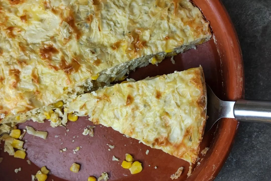 A baked savory pie with a golden, flaky crust is being served from a round brown dish; corn kernels and tender cabbage are visible in the filling, making it a delicious addition to your collection of comforting recipes.
