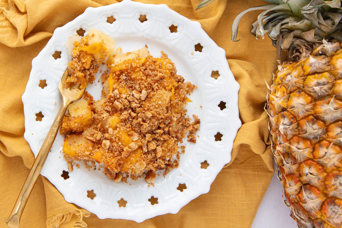 White plate with pineapple crumble dessert and a gold spoon on a yellow cloth, next to a whole pineapple—this sweet treat is inspired by unique casserole recipes you’ll want to save for later.