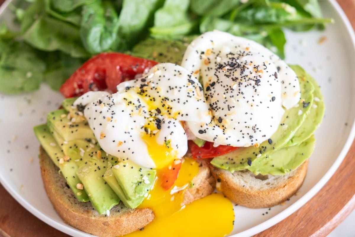 Two slices of bread topped with avocado, tomato, poached eggs, and poppy seeds, served with leafy greens on a white plate—a delicious twist on one of our most saved recipes.
