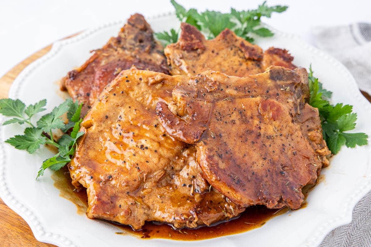 Three cooked pork chops with brown sauce, accented by maple syrup, served on a white plate and garnished with fresh parsley leaves—perfect for your collection of maple syrup recipes.