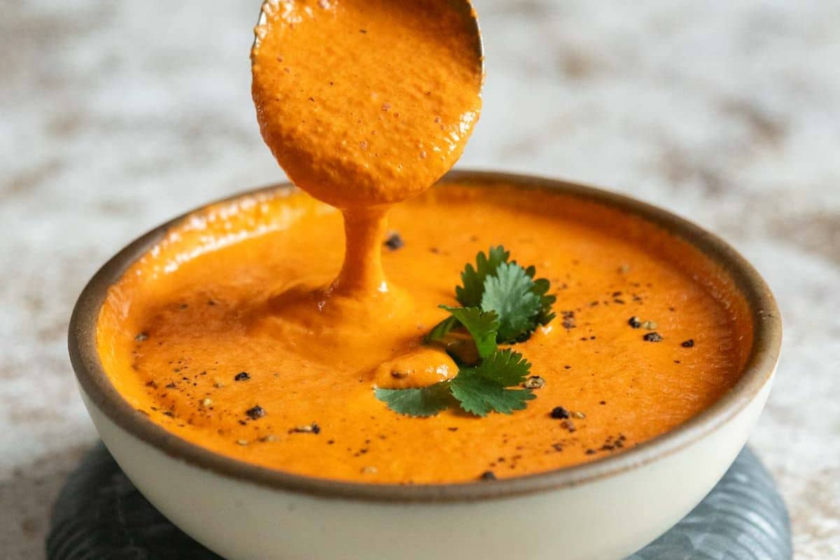 A spoon pours creamy orange sauce—made with roasted peppers—into a bowl topped with cilantro and black pepper.
