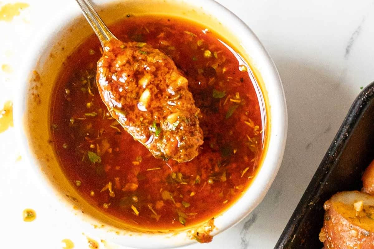 A spoon with red, oily sauce containing herbs and spices rests over a small white bowl filled with the same popular sauce on a white surface, perfect for adding bold flavor to your favorite recipes.