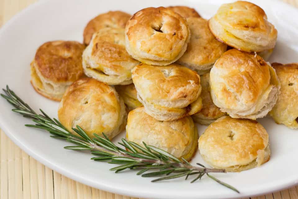 A plate of small, round, golden-brown puff pastry biscuits is garnished with a sprig of rosemary, making it a perfect addition to your collection of autumn recipes.