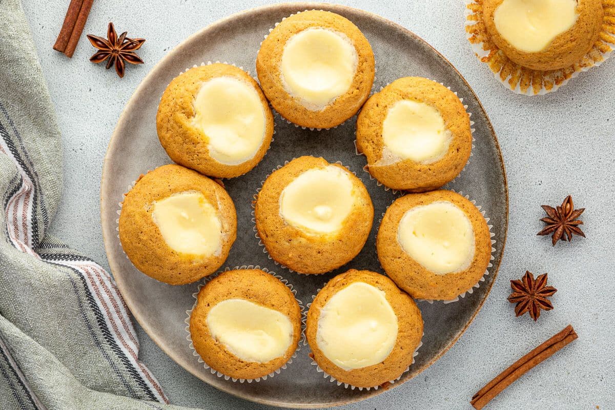 A plate of eight pumpkin muffins with cream cheese centers, perfect for make ahead recipes, sits on a light surface next to a striped cloth, cinnamon sticks, and star anise.