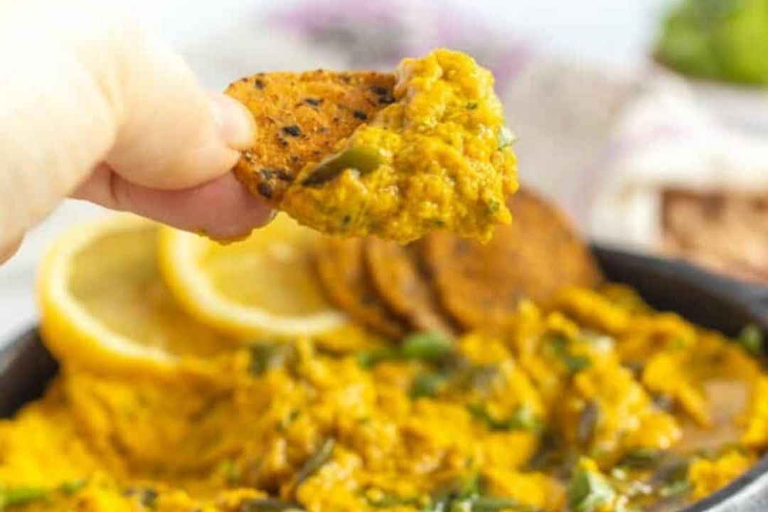 A hand dips a round cracker into a bowl of yellow dip with herbs, showcasing one of this season’s new pumpkin recipes; lemon slices and more crackers are visible in the background.