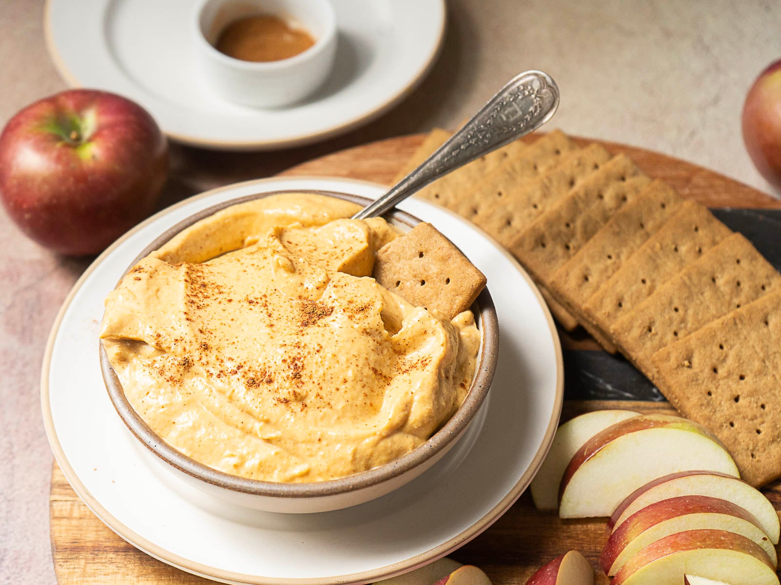 A bowl of creamy dip with a cracker, surrounded by graham crackers, apple slices, and a whole apple—perfect for maple syrup recipes or creative maple syrup uses.