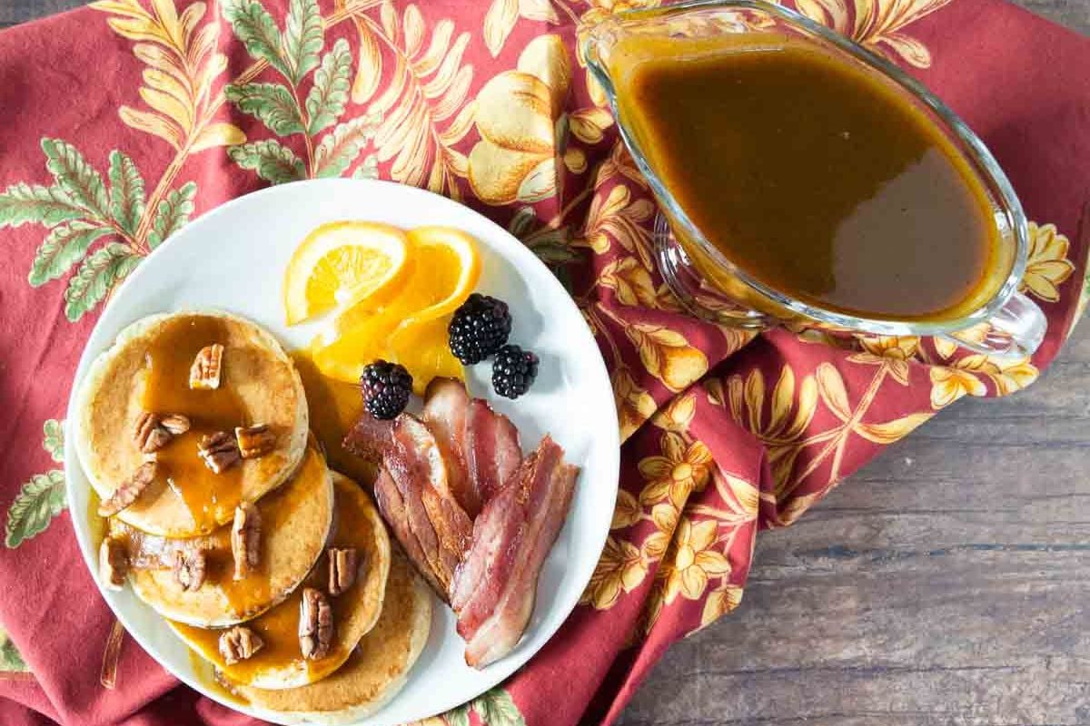 A plate with pancakes topped with sauce and pecans, crispy bacon, orange slices, and blackberries sits alongside a gravy boat of brown sauce on a red patterned napkin—perfect for savoring cozy autumn mornings or trying out delicious new pumpkin recipes.