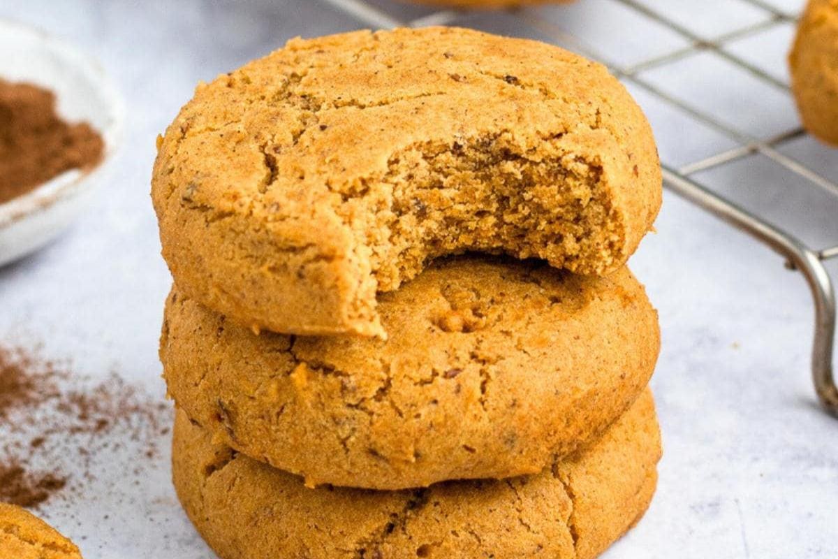 Three stacked cookies, with the top cookie missing a bite, sit on a white surface next to a cooling rack and a small bowl of cocoa powder—perfect for kids and easy desserts that capture the cozy spirit of Autumn.