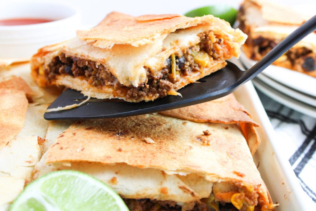 A slice of baked quesadilla with ground beef and vegetables is being lifted with a spatula from a tray—an ideal option for easy dinners or sheet pan meals. A lime wedge and sauce are visible nearby.