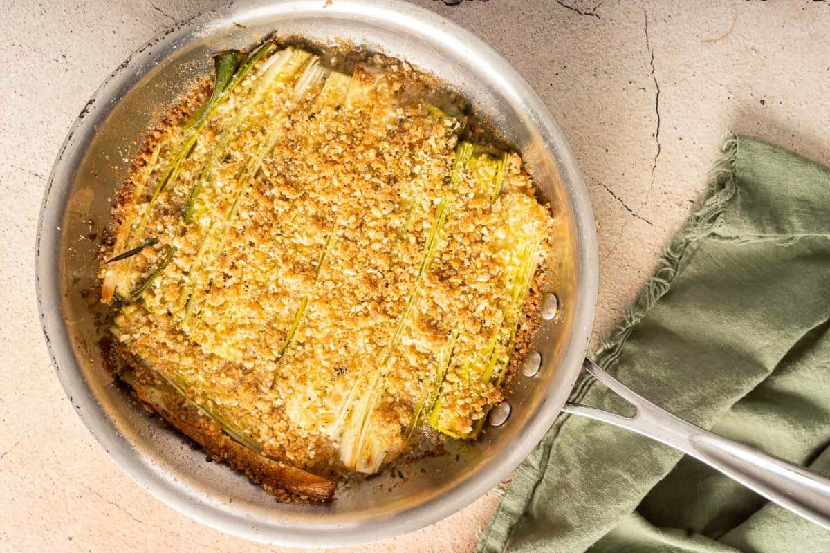 A pan of baked leeks topped with a golden breadcrumb crust sits on a light textured surface, perfect for fans of roast vegetables, next to a green cloth napkin.
