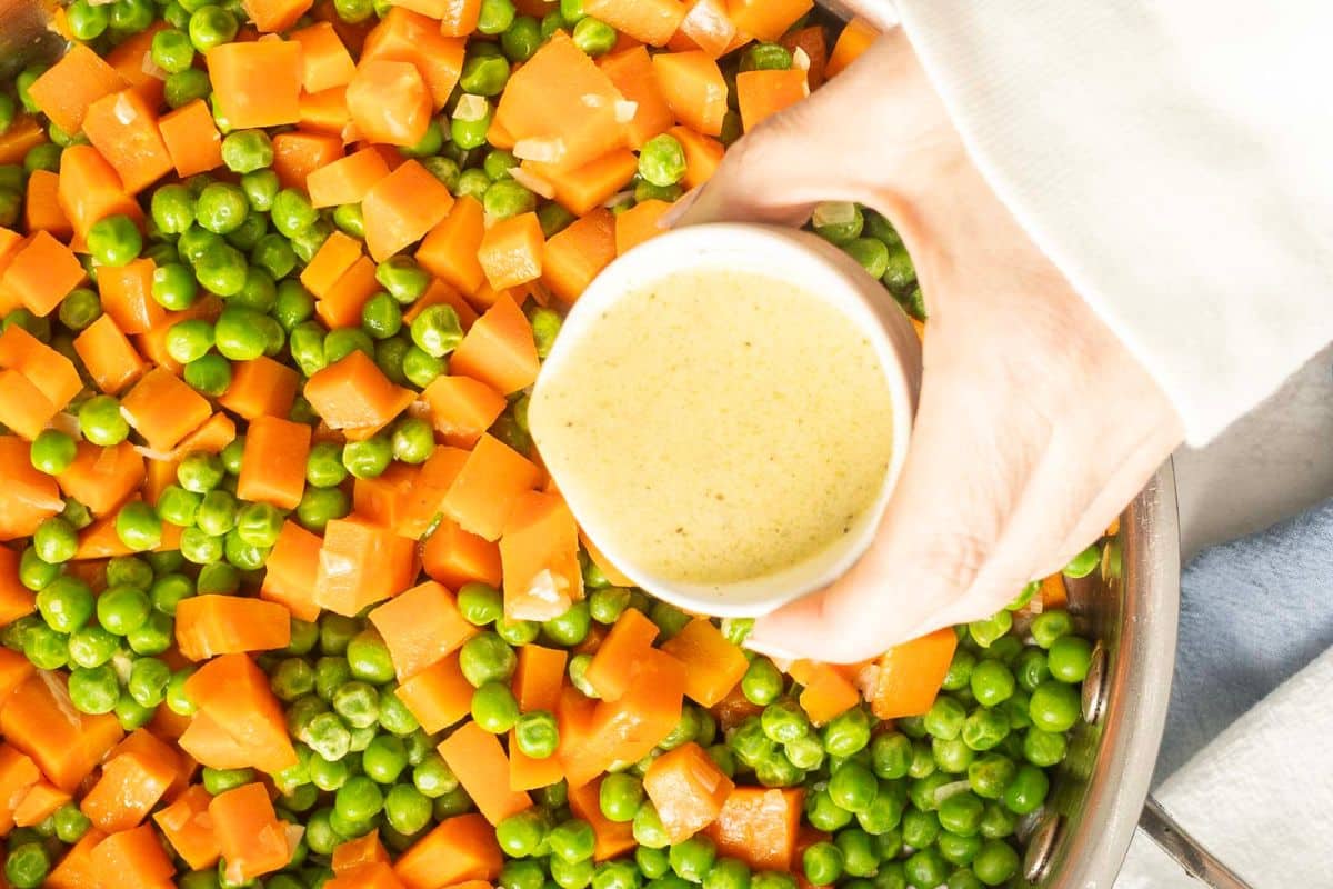 A hand holds a small bowl of sauce above a pan filled with cooked diced carrots and green peas—perfect for easy carrot recipes or busy weeknight sides.