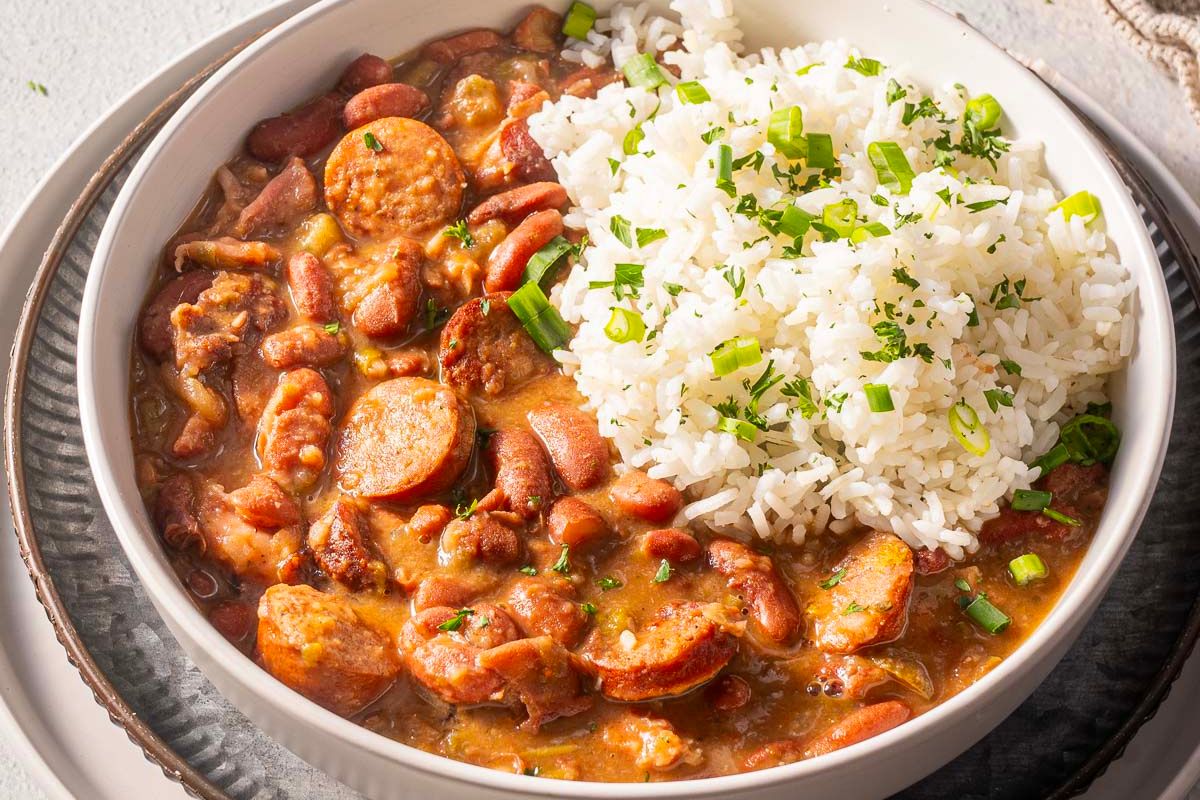 A bowl of white rice garnished with chopped green onions is served next to red beans and sausage in a creamy sauce—an evergreen dish inspired by timeless recipes.
