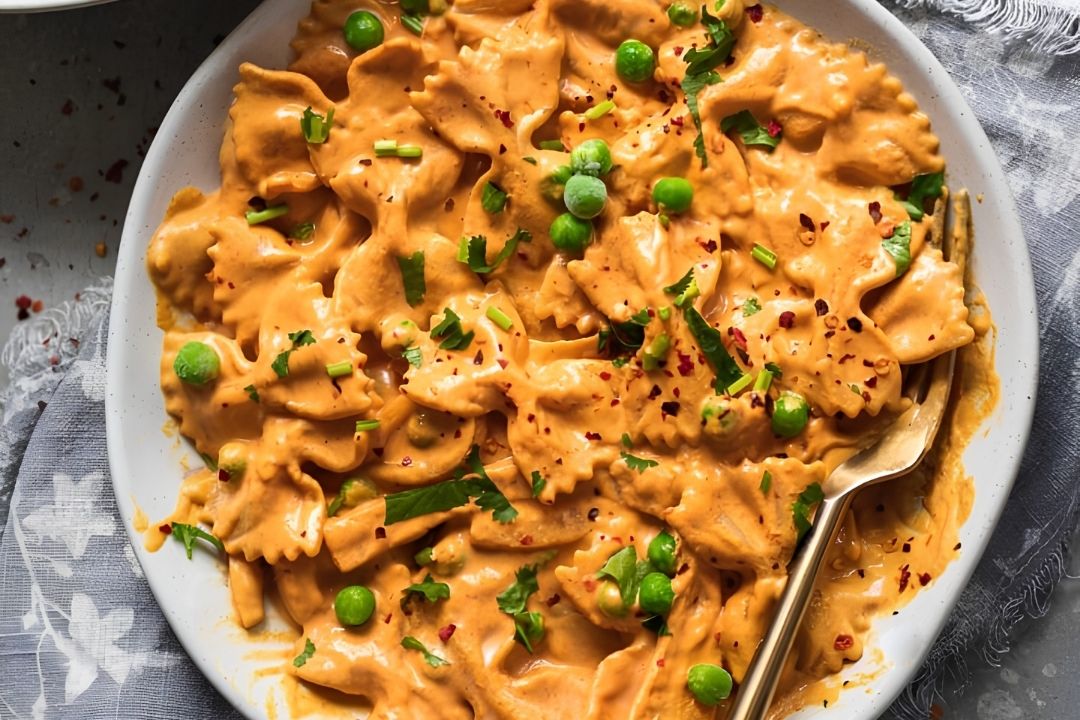 A plate of bowtie pasta in a creamy orange sauce, topped with green peas, chopped herbs, and red pepper flakes—perfect for anyone searching for delicious recipes with roasted peppers. A fork sits invitingly on the side.