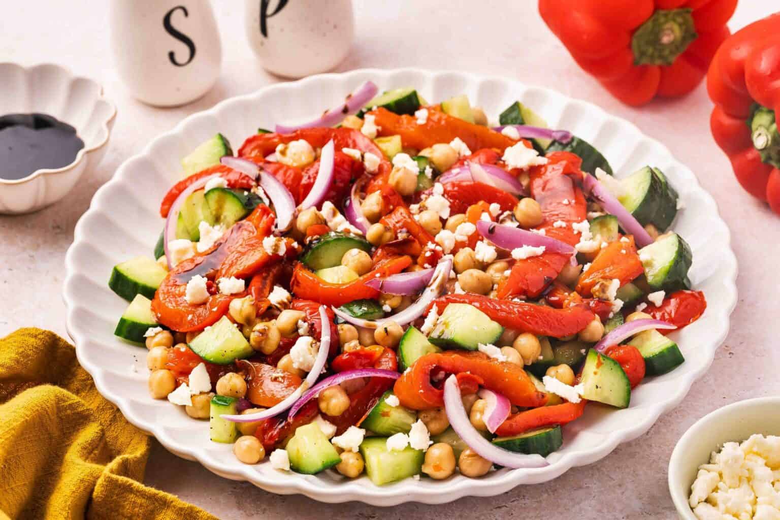 A plate of salad with cucumbers, chickpeas, roasted red peppers, red onions, and feta cheese sits invitingly on the table, perfect inspiration for anyone looking for delicious recipes with roasted peppers. Salt and pepper shakers and a jar of roasted peppers add to the colorful scene, while fresh red bell peppers rest in the background.