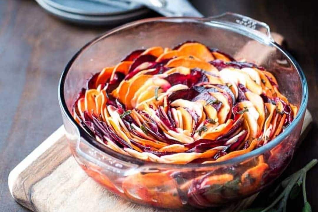 A glass baking dish filled with thinly sliced, layered root vegetables arranged in a circular pattern, resting on a wooden board—perfect for cozy autumn meals and inspired by Grandma-approved recipes.