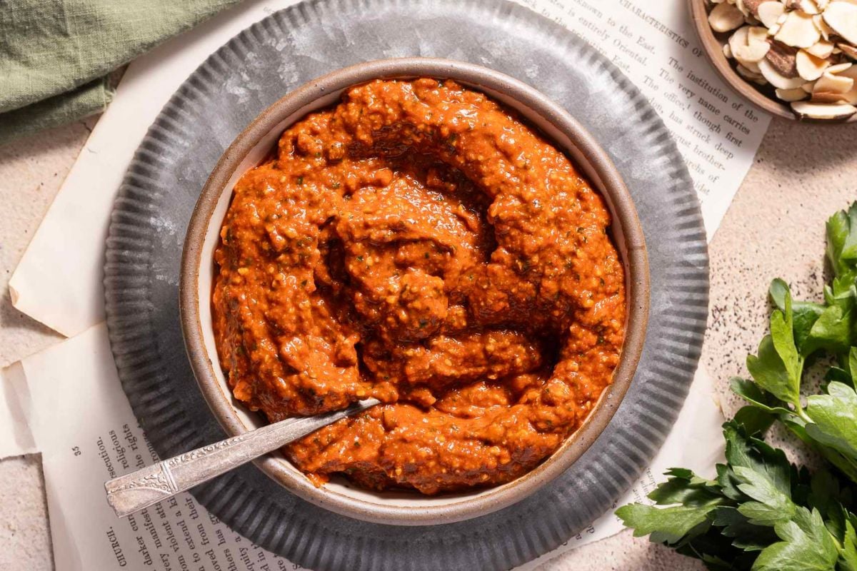 A bowl of red romesco sauce with a spoon rests on a plate, surrounded by parsley, almonds, and pages of a book—offering inspiration for recipes with roasted peppers.