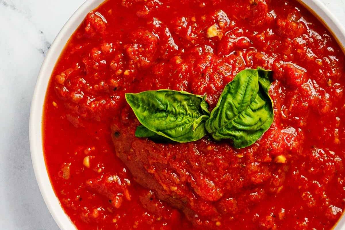 Bowl of tomato sauce topped with two fresh basil leaves, viewed from above on a white surface—a staple in classic recipes and traditional dishes.