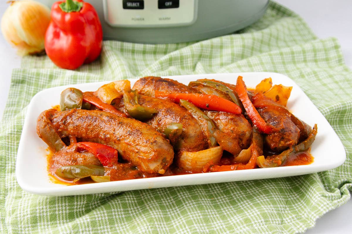 A rectangular white plate with sausages, chicken, bell peppers, and onions in tomato sauce sits on a green plaid cloth, offering a flavor-packed dinner option, with a slow cooker in the background.