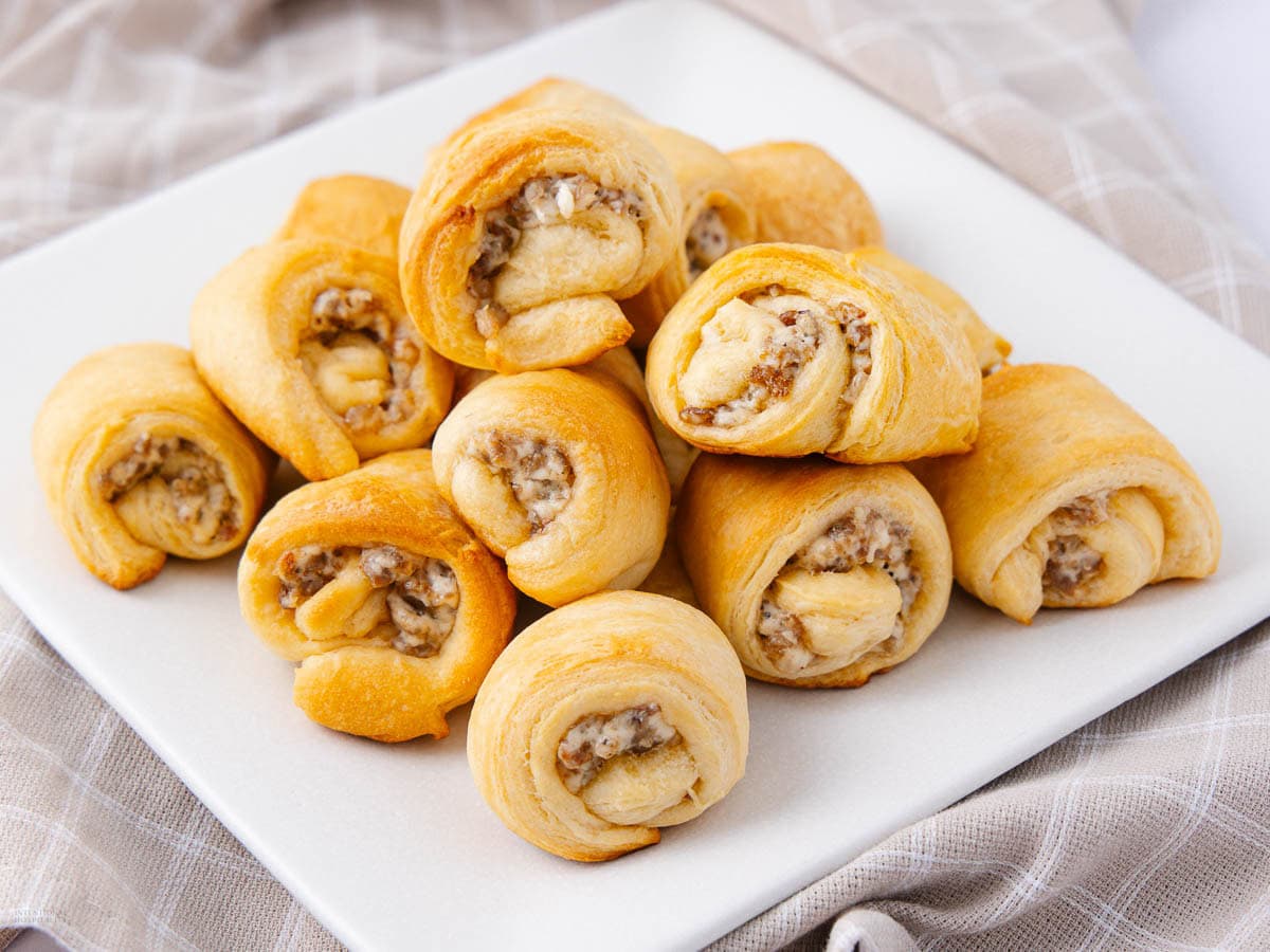 A white plate holds several golden-brown sausage rolls with visible filling, arranged in a pile on a light plaid cloth—perfect for easy recipes or week meal prep.