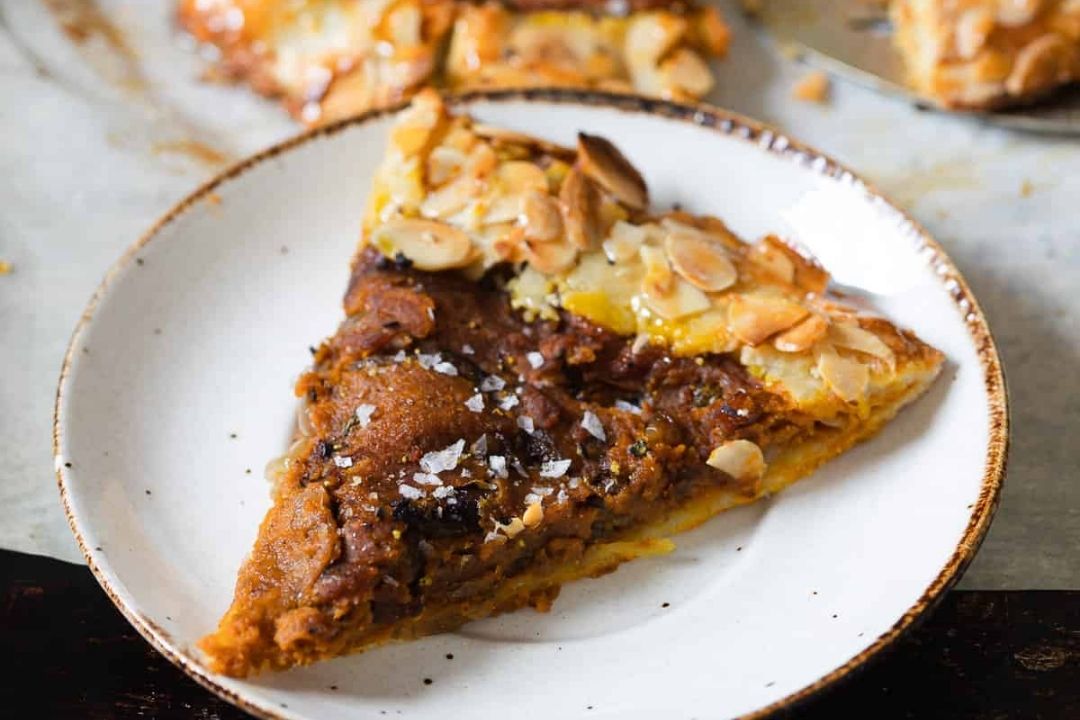 A slice of pie with a dark filling, topped with sliced almonds and coarse salt, sits on a white plate with a brown rim—an irresistible treat inspired by grandma-approved recipes and perfect for autumn dishes.