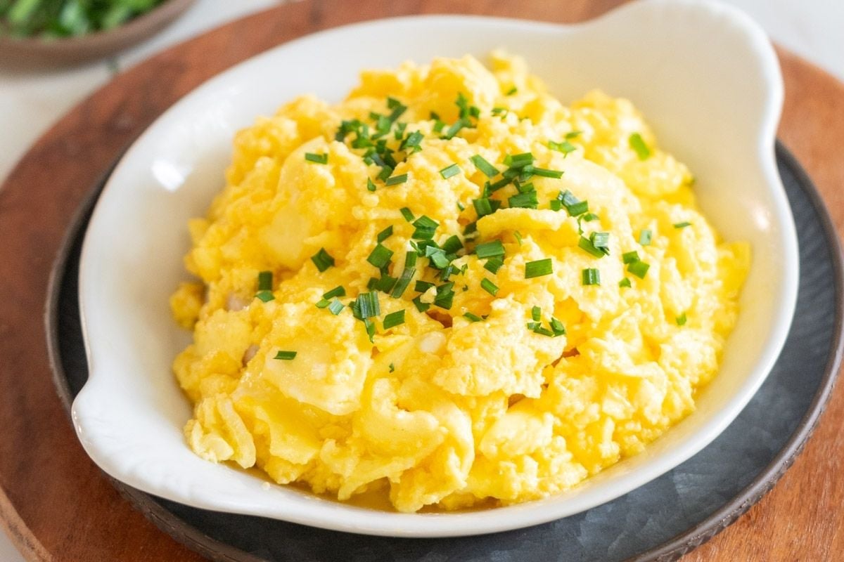 A white dish filled with scrambled eggs garnished with chopped chives, placed on a wooden surface—one of the classic recipes everyone should learn.