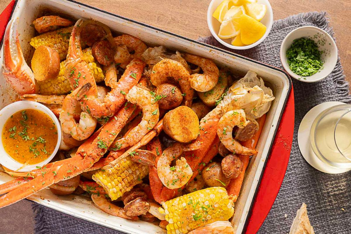 A tray of Southern Supers seafood boil with crab legs, shrimp, sausage, corn, and potatoes, served with lemon wedges, chopped herbs, dipping sauce, and a glass of white wine on the side.