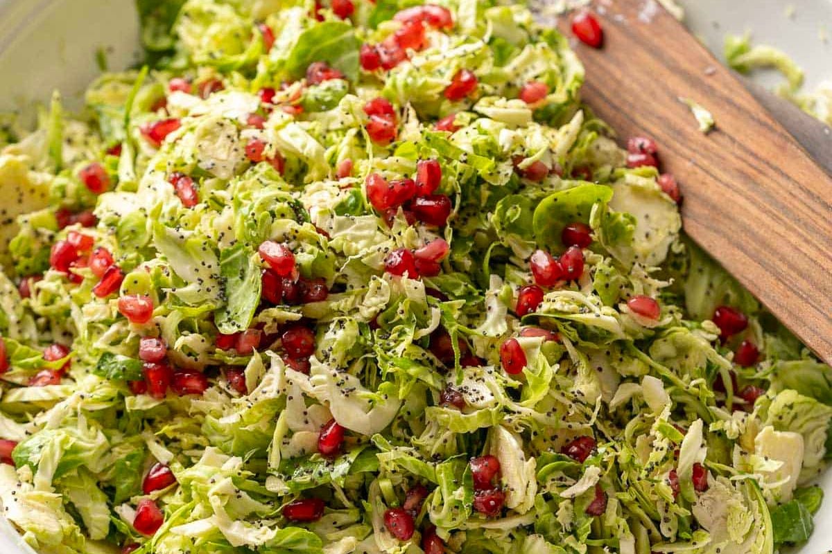 Shredded Brussels sprouts salad topped with pomegranate seeds and cracked black pepper, perfect for salad season and delicious as part of your favorite autumn recipes, with a wooden spoon resting on the side.