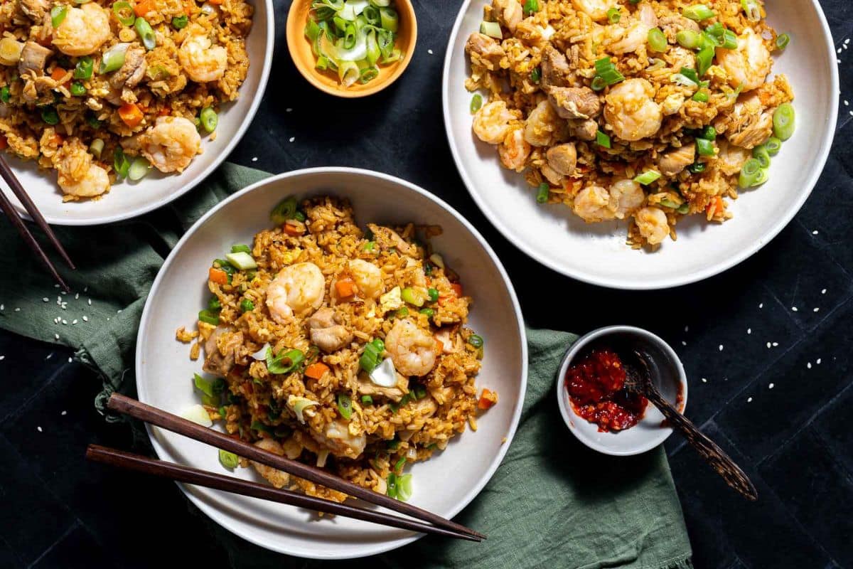 Three bowls of flavorful rice with shrimp, chicken, vegetables, and green onions are served with chopsticks, chili sauce, and a small bowl of sliced scallions on a dark table.