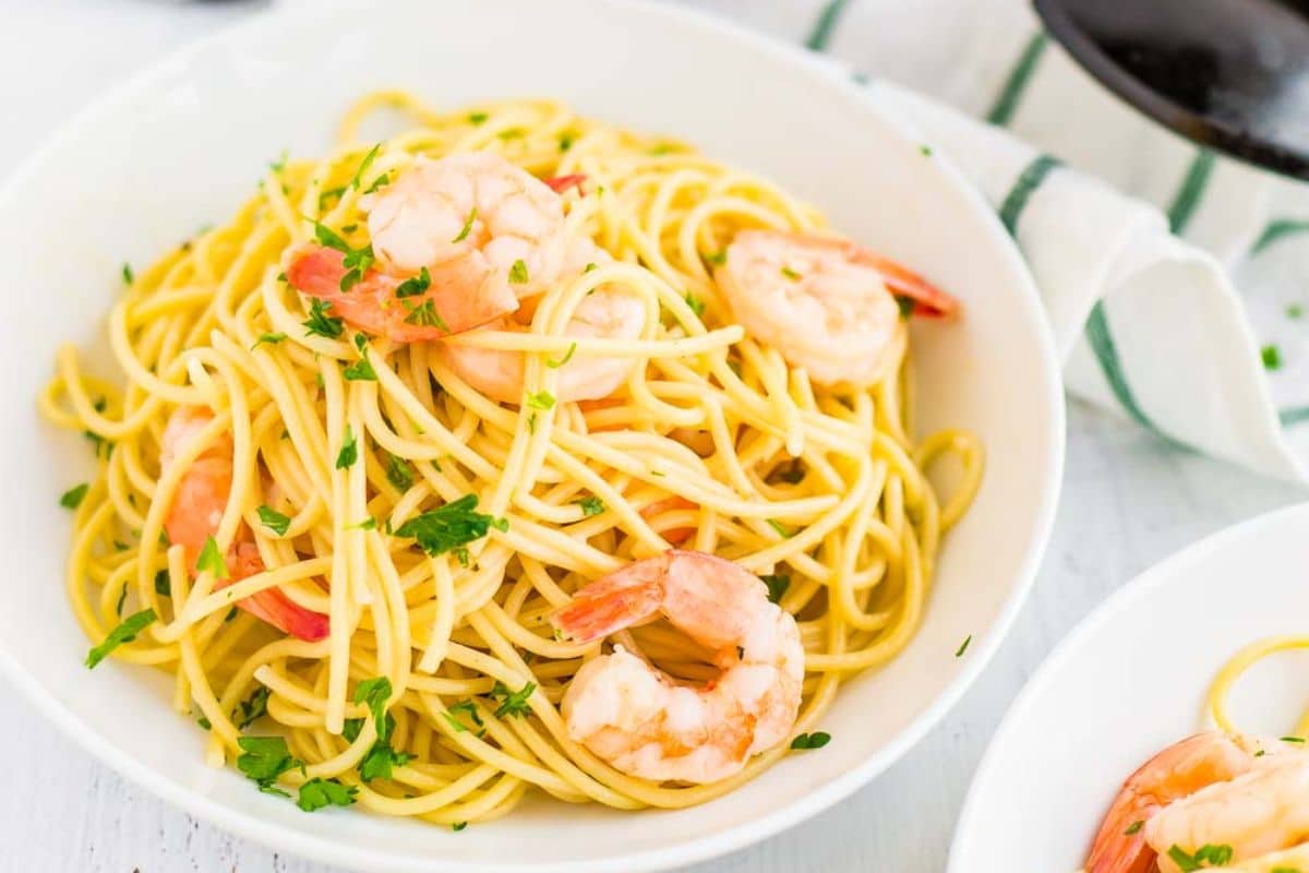 A white bowl filled with spaghetti and shrimp, garnished with chopped parsley, on a white table with a striped napkin nearby—one of those essential recipes every home cook should learn.