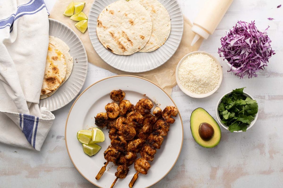 Grilled shrimp skewers on a plate with lime wedges, surrounded by tortillas, avocado, cilantro, shredded cabbage, cheese, and a bottle of sauce on a white surface—perfect for those seeking fresh Tex Mex dinner ideas.