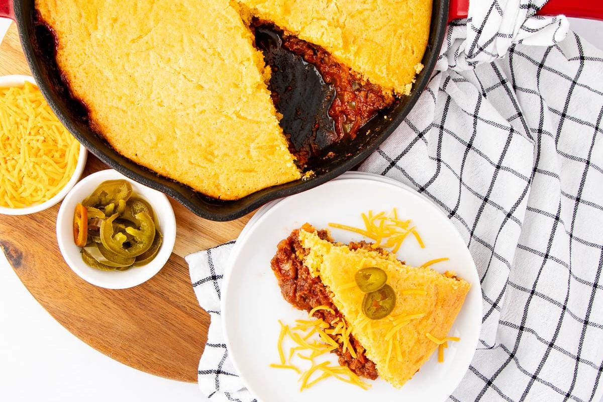 A slice of cornbread-topped chili pie on a plate, garnished with jalapeños and shredded cheese, sits next to a skillet filled with the remaining pie, cheese, and jalapeños—one of those unique casserole recipes that’s perfect for anyone seeking new casserole ideas.