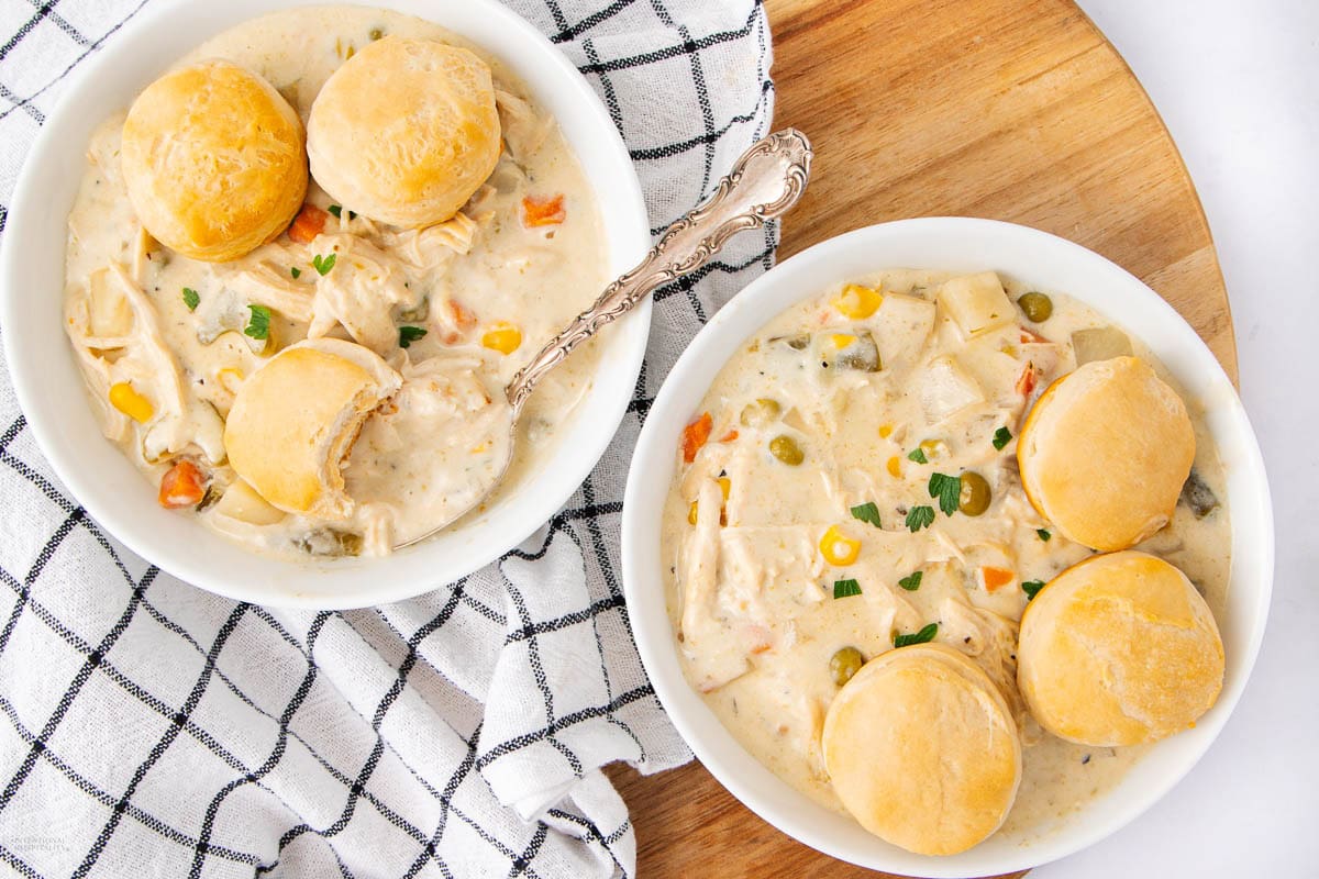 Two bowls of creamy chicken and vegetable stew made with rotisserie chicken, each topped with biscuits, sit on a wooden board next to a black and white checkered towel.
