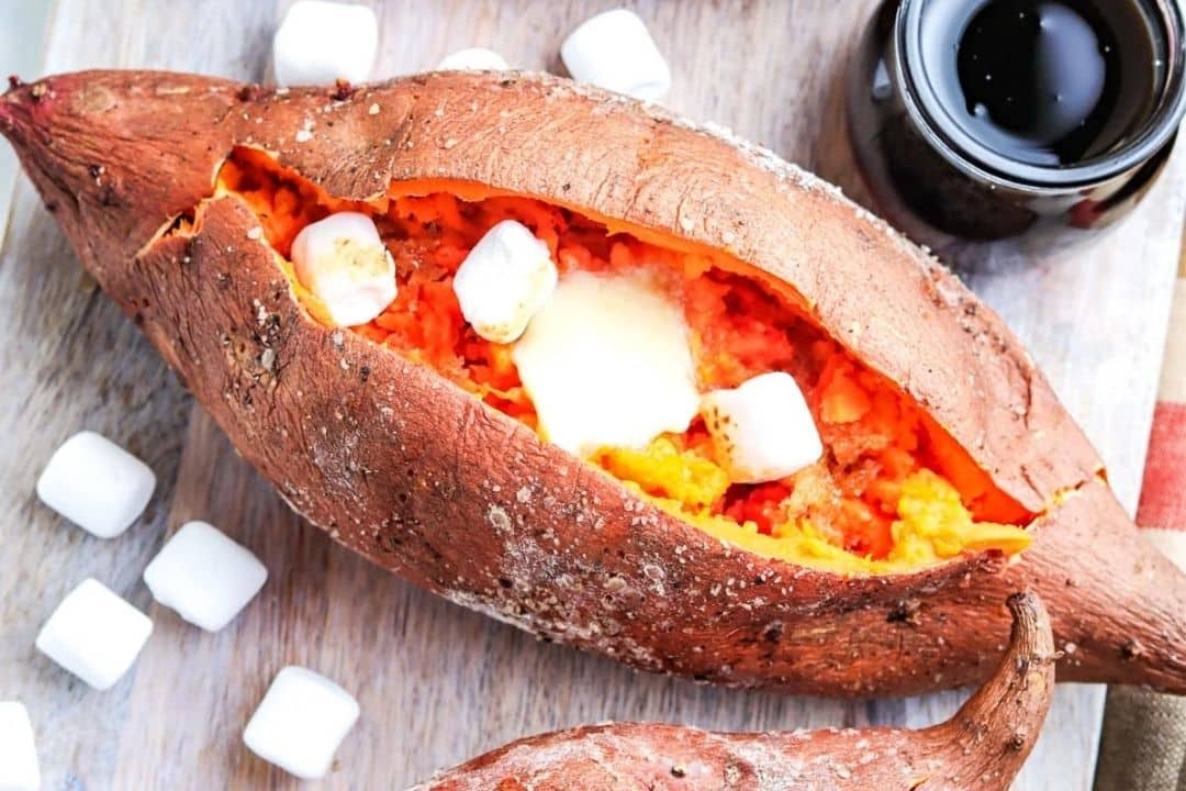 Baked sweet potato split open, filled with butter and mini marshmallows—a cozy classic among sweet potato recipes—on a wooden surface with extra marshmallows and a cup of dark liquid.