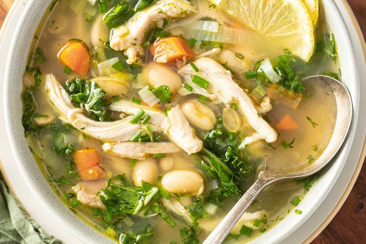 Bowl of chicken and white bean soup with carrots, spinach, herbs, and a lemon slice, with a spoon resting inside—perfect for easy chicken recipes or cozy weeknight dinners.