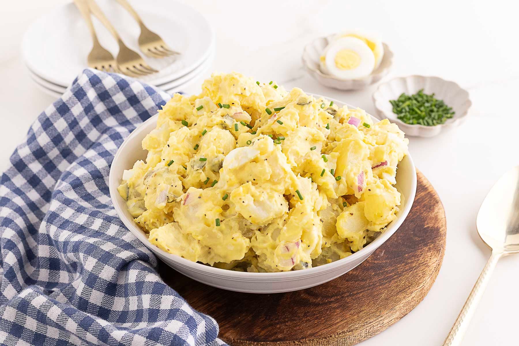 A bowl of potato salad garnished with chopped chives sits on a wooden board beside a blue checkered napkin, plates, forks, and a sliced hard-boiled egg—a stress free dish that’s perfect to feed a crowd at your next gathering or as one of your go-to potluck recipes.