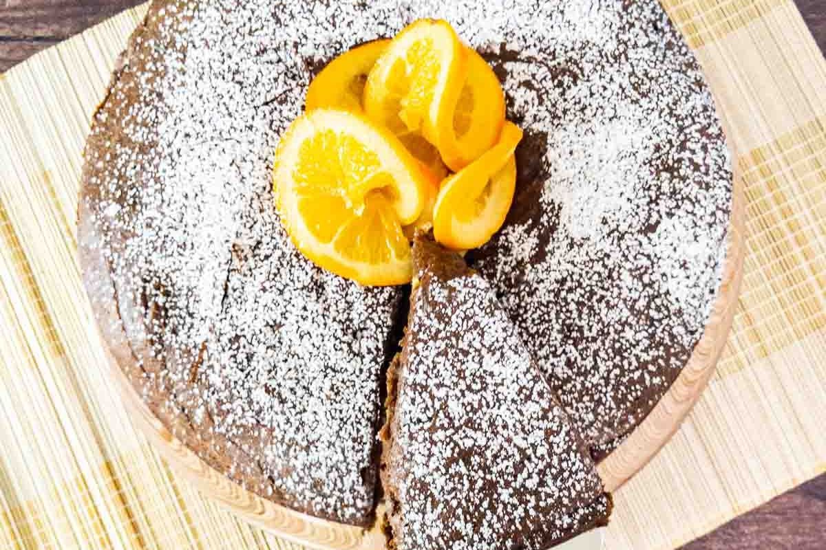 Chocolate cake topped with powdered sugar and orange slices, with one slice partially removed, placed on a woven mat—one of our Fall Favorites that’s also gluten free.