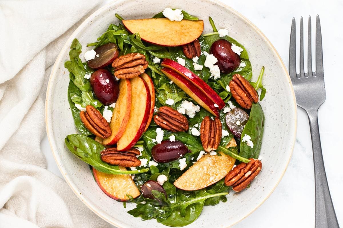 A bowl of salad with spinach, arugula, sliced peaches, grapes, pecans, and crumbled feta cheese next to a fork—perfect for celebrating salad season.