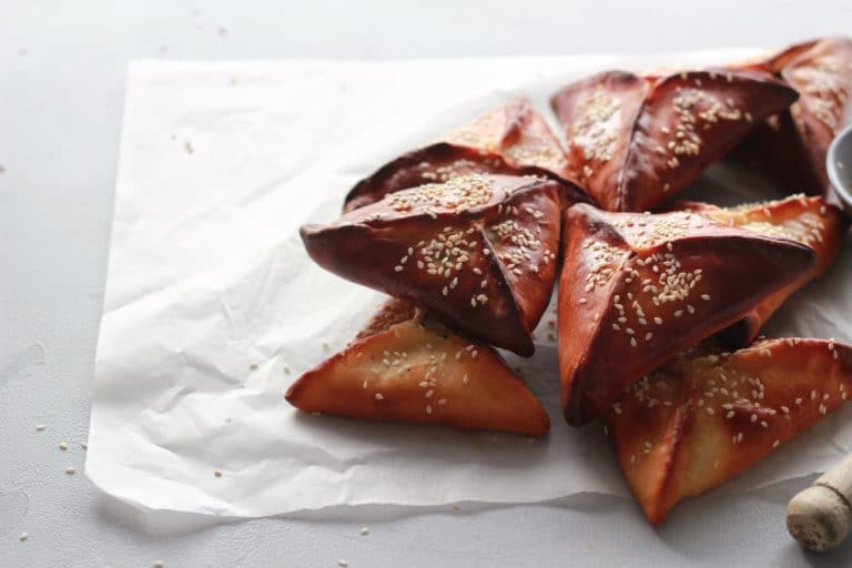 Six triangular pastries topped with sesame seeds are arranged on a sheet of white parchment paper, with part of a rolling pin visible in the corner—an inviting addition to your collection of savory breakfast recipes.