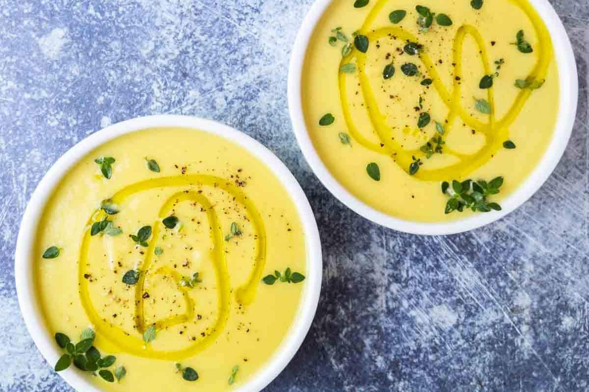 Two white bowls filled with yellow soup, garnished with fresh herbs, black pepper, and a drizzle of olive oil, placed on a textured gray surface—perfect inspiration for your saved recipes or to add to your collection of popular recipes.